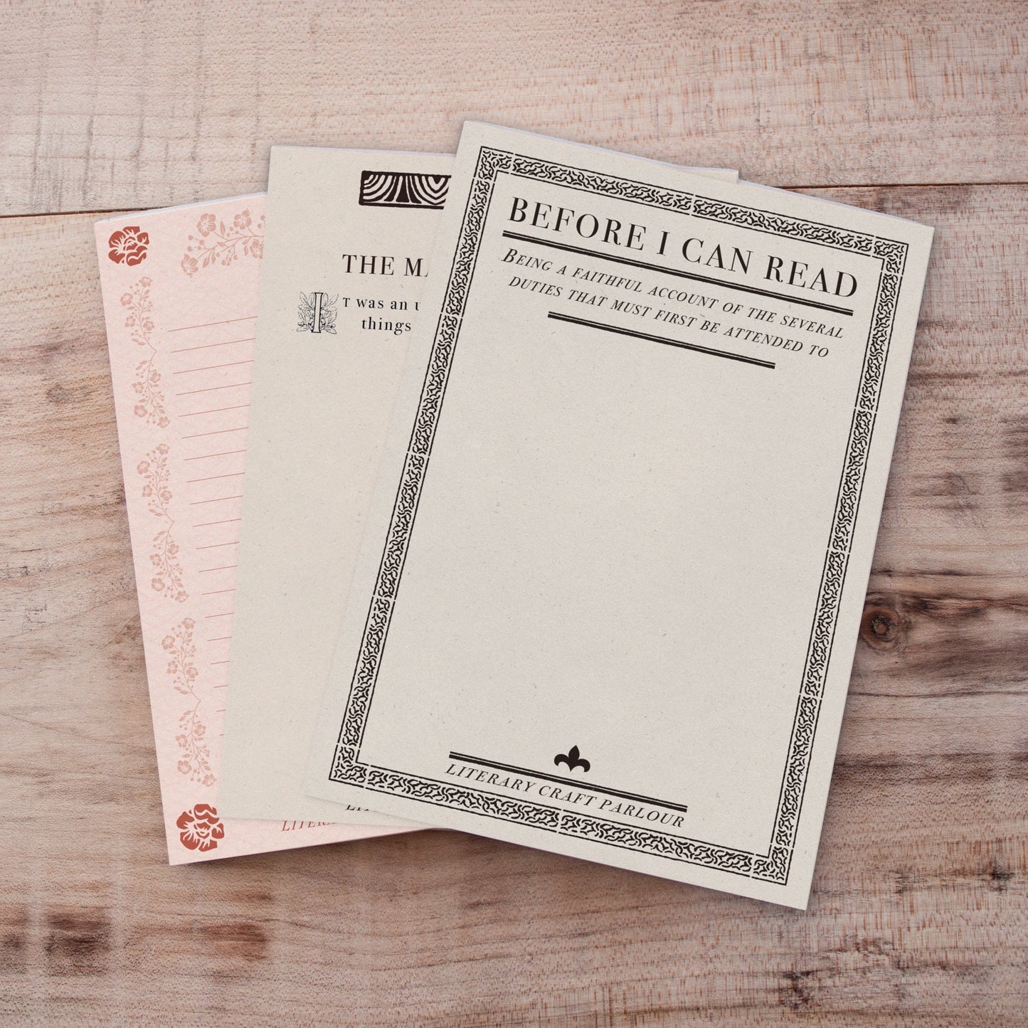 Three vintage-style book-themed notepads stacked on a light wood background. The top card is cream with a black decorative border and the text 'BEFORE I CAN READ'.