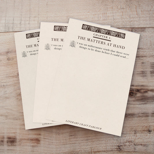 Three cream-colored book page notepads fanned out on a light wood surface, printed with black decorative text and geometric borders.