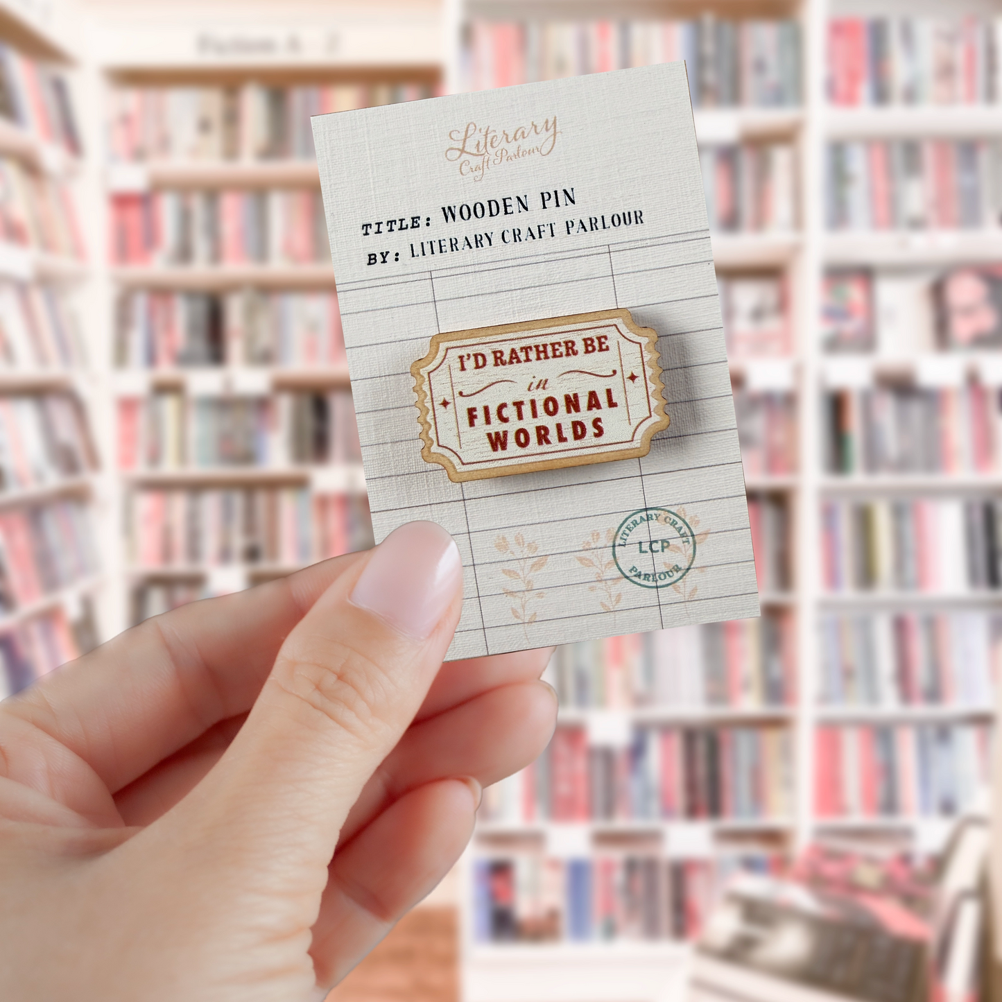 A hand holds a small wooden pin mounted on a branded backing card. The pin is shaped like a vintage ticket with the text 'I'D RATHER BE IN FICTIONAL WORLDS' in red lettering.
