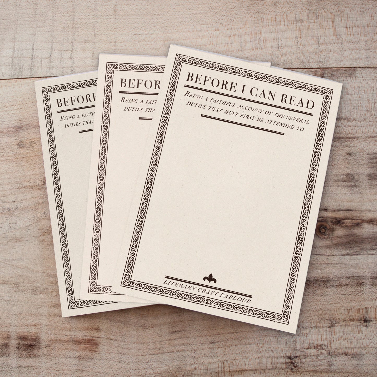 Three vintage-style notepads fanned out on a light wood background. The notepads are off-white with ornate dark borders and the title 'BEFORE I CAN READ' printed at the top.