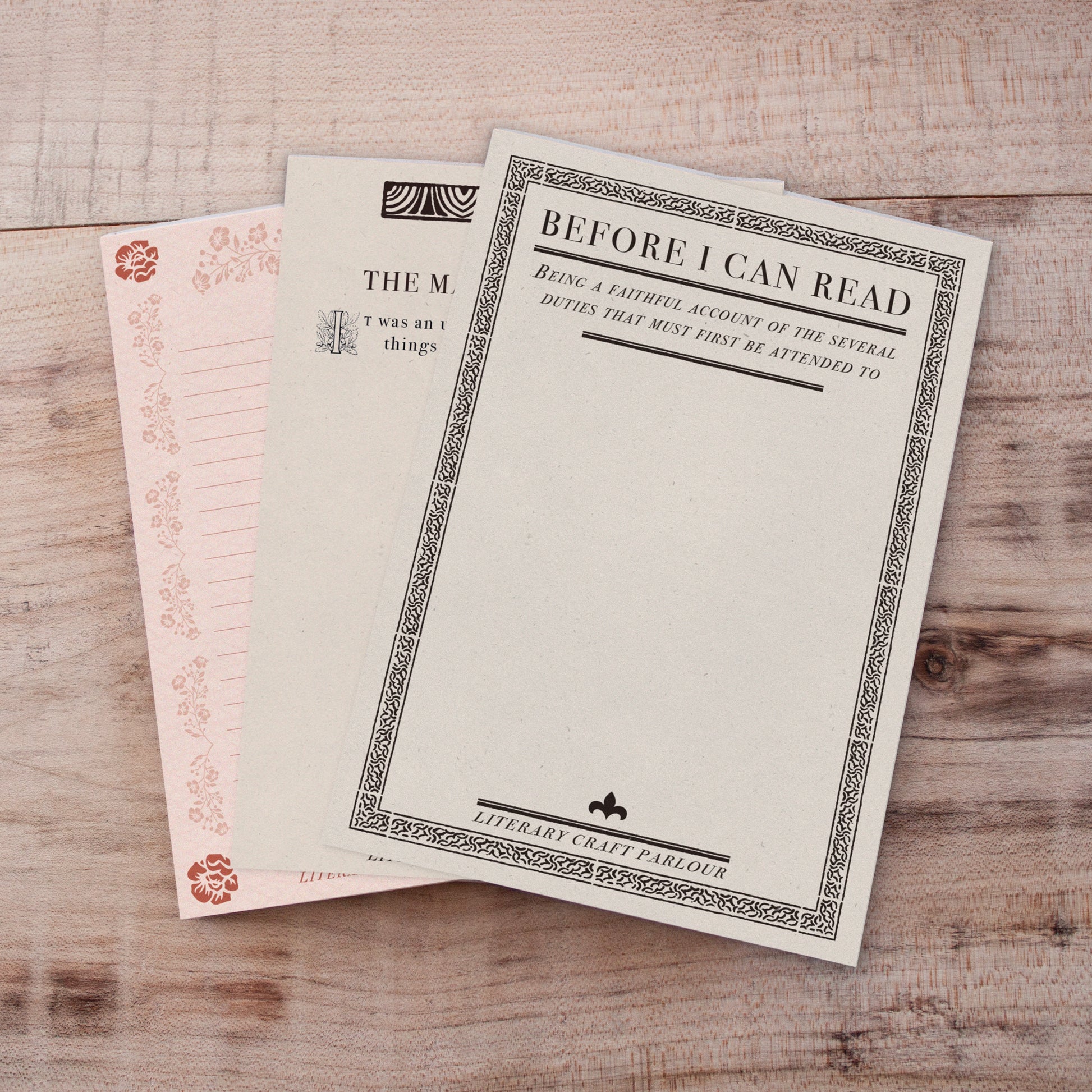 Three vintage-style book-themed notepads stacked on a light wood background. The top card is cream with a black decorative border and the text 'BEFORE I CAN READ'.