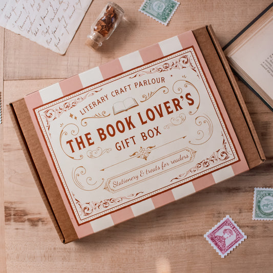A flat-lay view of 'The Book Lover's Gift Box' by Literary Craft Parlour, placed on a wooden table with vintage props like old books, stamps, and a letter. The box has a decorative pink and white striped background with red text and red and gold borders and accents. The style is similar to a vintage chocolate to cookie box or tin.
