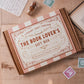 A flat-lay view of 'The Book Lover's Gift Box' by Literary Craft Parlour, placed on a wooden table with vintage props like old books, stamps, and a letter. The box has a decorative pink and white striped background with red text and red and gold borders and accents. The style is similar to a vintage chocolate to cookie box or tin.