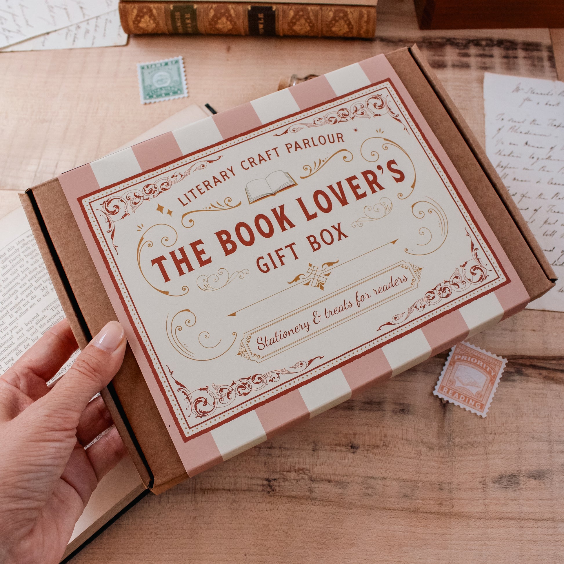 A hand holds a brown cardboard gift box labeled 'The Book Lover's Gift Box' with a pink and cream striped ribbon. The box is photographed on a wooden tabletop with vintage-style stationery props around it.