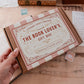 A hand holds a brown cardboard gift box labeled 'The Book Lover's Gift Box' with a pink and cream striped ribbon. The box is photographed on a wooden tabletop with vintage-style stationery props around it.