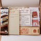 A curated vintage-themed reading gift set displayed inside a cardboard box, featuring a Galaxy instant chocolate sachet, a task list notepad, a reading list bookmark, a wooden book pin, a vintage-style book stamp, and a book club ticket.
