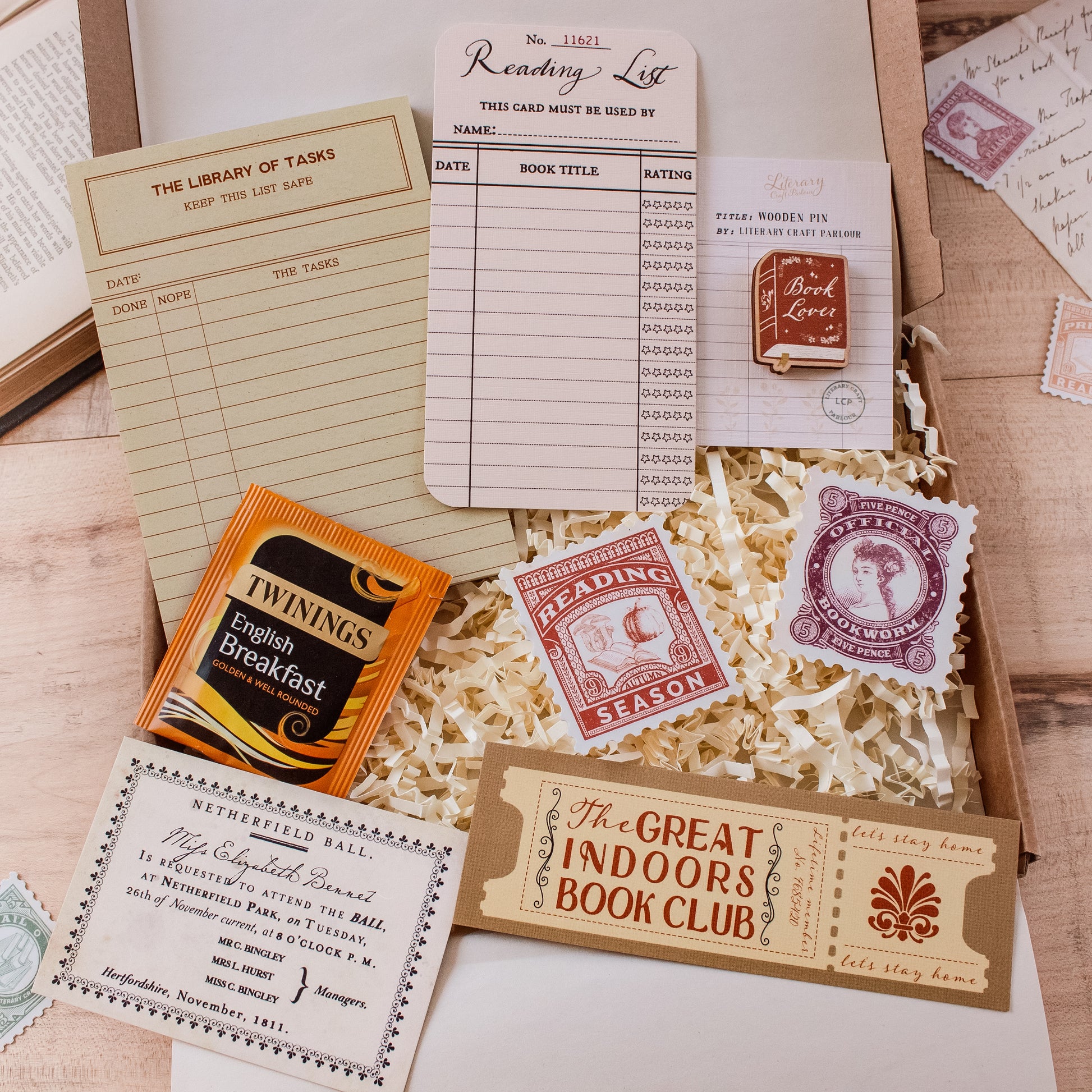 A styled flat-lay of vintage-inspired reading and library-themed stationery items arranged in shredded paper inside an open box. Visible items include a reading list card, library of tasks to do-list notepad, a wooden book lover pin badge, reading season and official bookworm stamp-shaped stickers, ticket-style bookmarks, and a Twinings English Breakfast tea packet.