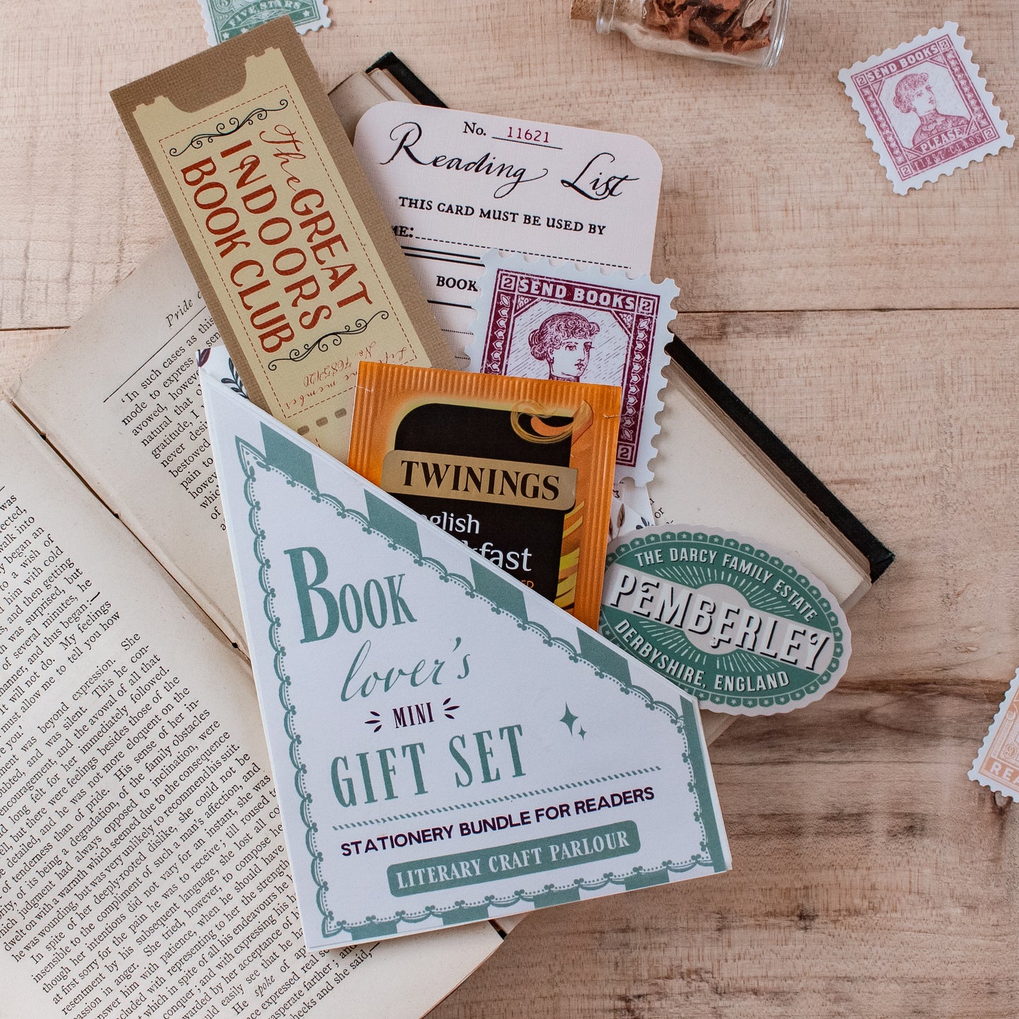 A stationery gift set that says 'Book lover's mini GIFT SET'. A stationery bundle for readers styled on a wooden tabletop with an open book. The set includes a ticket style bookmark, a reading list card, a vintage-style stamp sticker reading "send books please", a Pemberley vintage travel sticker, and a Twinings tea packet tucked inside.