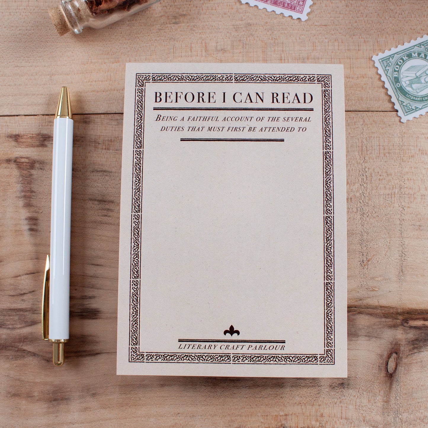 A cream-coloured notepad designed to look like a vintage book title page. It has a vintage-style black border and the heading text 'BEFORE I CAN READ' with a subheading "Being a faithful account of the several duties that must first be attended to" then leaving blank space for the user to write their own lists. The notepad lies on a wooden tabletop, styled with a white pen with gold trim, vintage postage stamps, and a small glass vial.