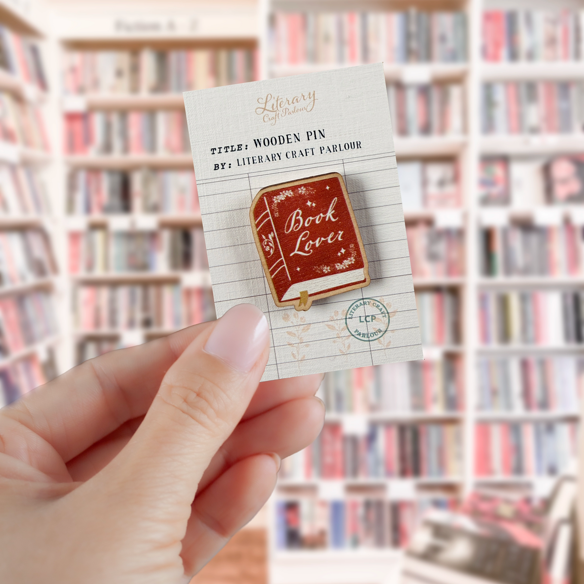 A hand holds a small printed card featuring a wooden pin shaped like a book with the words 'Book Lover' on the cover. The background is a softly blurred library bookshelf scene.