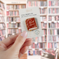 A hand holds a small printed card featuring a wooden pin shaped like a book with the words 'Book Lover' on the cover. The background is a softly blurred library bookshelf scene.
