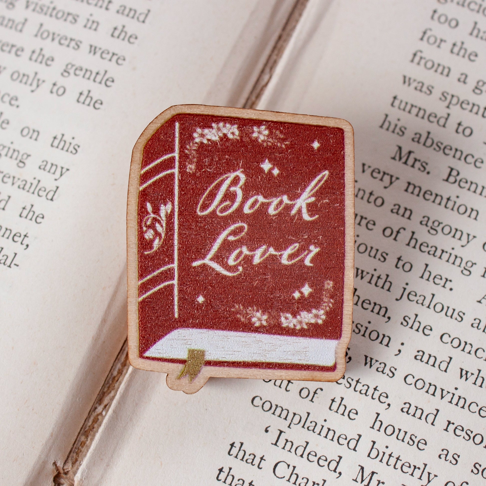 A rectangular book-shaped pin with a red cover reading 'Book Lover' in white script, placed on an open book page.