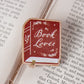 A rectangular book-shaped pin with a red cover reading 'Book Lover' in white script, placed on an open book page.