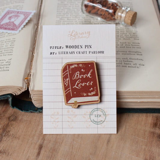A wooden book lover bookmark pin with 'Book Lover' written on it, mounted on a white card labeled 'Literary Craft Parlour' and placed on an open book over a wooden surface.
