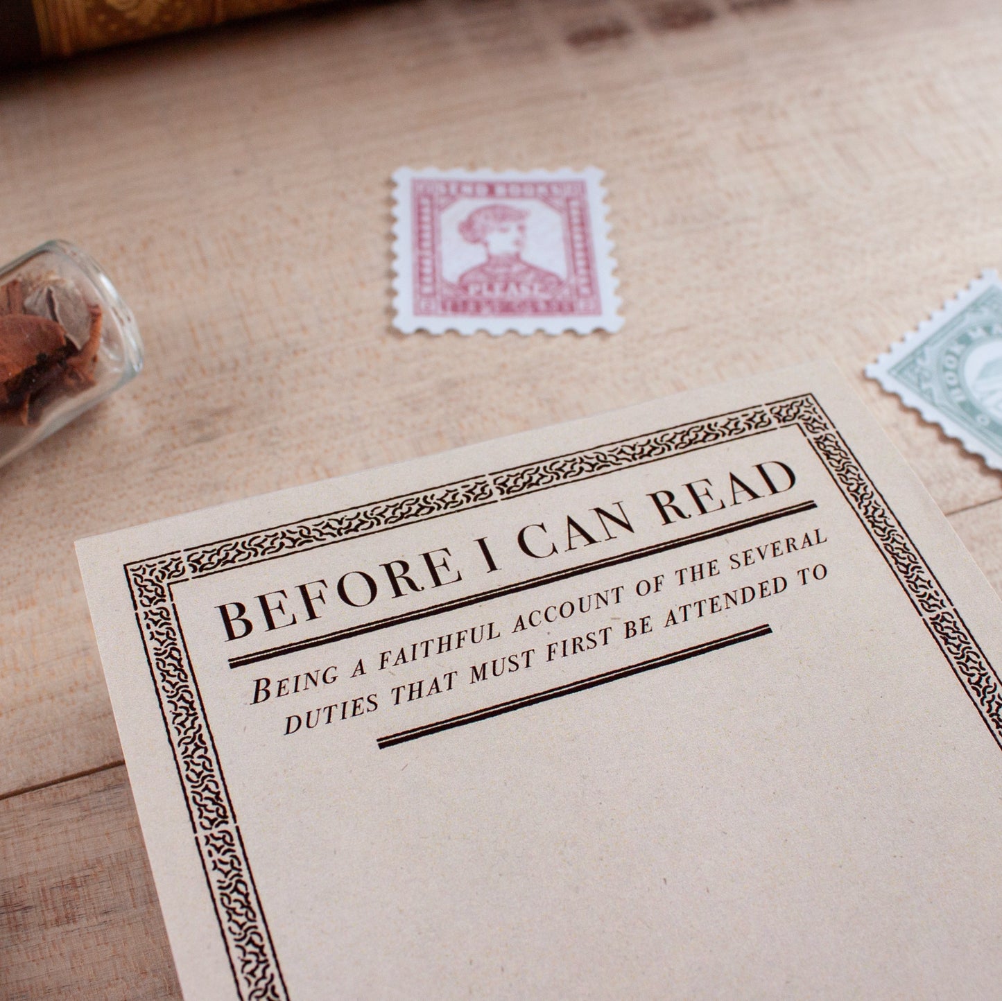A vintage-style notepad titled 'BEFORE I CAN READ' with decorative black border text on cream paper, styled on a pale wooden surface with old postage stamps nearby.