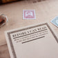 A vintage-style notepad titled 'BEFORE I CAN READ' with decorative black border text on cream paper, styled on a pale wooden surface with old postage stamps nearby.