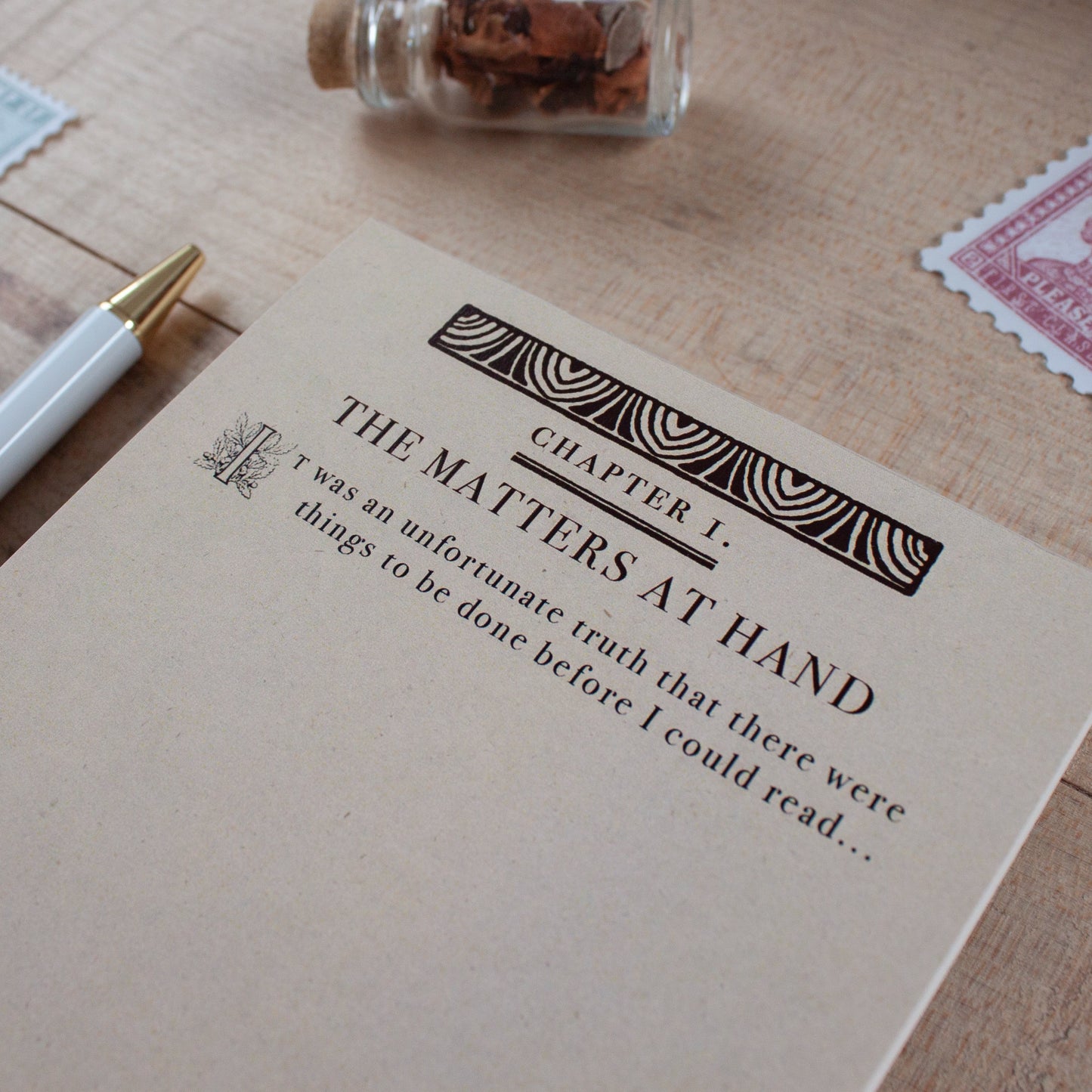A close-up of a notepad page titled "THE MATTERS AT HAND" with decorative typography, placed on a light wood surface with a white pen, a small glass bottle, and postage-style stamps nearby.