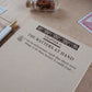 A close-up of a notepad page titled "THE MATTERS AT HAND" with decorative typography, placed on a light wood surface with a white pen, a small glass bottle, and postage-style stamps nearby.