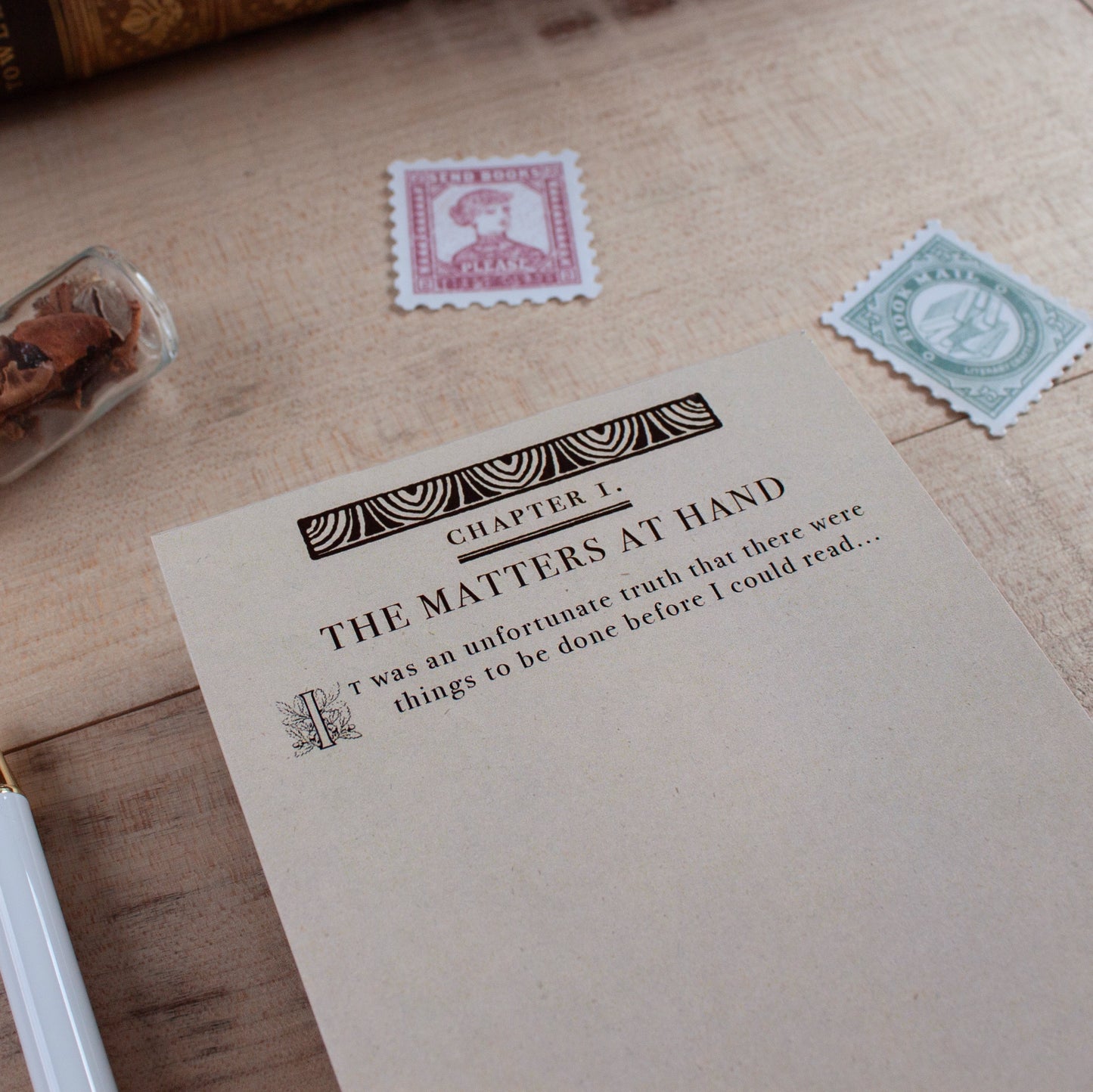 A vintage-style notepad with the heading 'Chapter 1. The Matters at Hand'. The notepad is designed to look like the first page of a vintage book and starts with the quote "It was an unfortunate truth that there were things to be done before I could read..." then leaving blank space for the user to write their own to-do list making it seem like their to-do list is the start of an epic tale. The notepad is placed on a wooden surface with old postage stamps nearby.