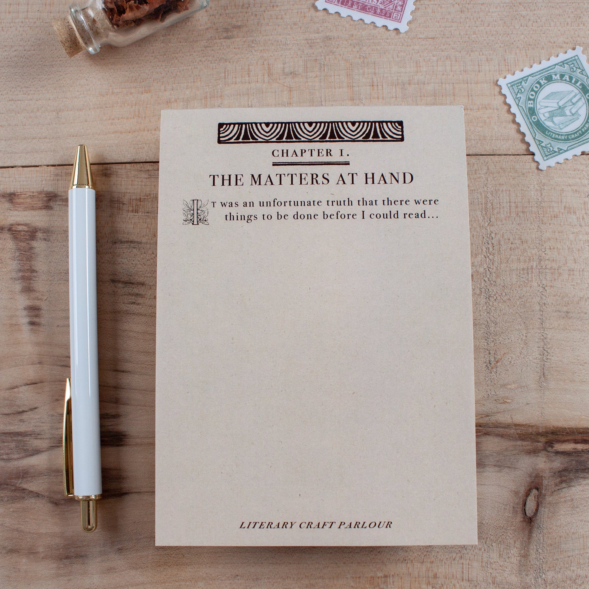 A styled flat-lay of a notepad titled 'Chapter 1. THE MATTERS AT HAND' on a wooden tabletop. The notepad is designed to look like the first page of a vintage book and starts with the quote "It was an unfortunate truth that there were things to be done before I could read..." then leaving blank space for the user to write their own to-do list making it seem like their to-do list is the start of an epic tale. It is styled on a wooden desk with a white and gold pen and  vintage postage stamps.
