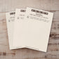 Three cream-colored book page notepads fanned out on a light wood surface, printed with black decorative text and geometric borders.