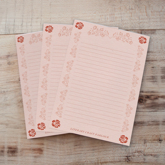 Three pink lined notepads with a rose gold floral border and corner accents, arranged on a rustic wooden surface.