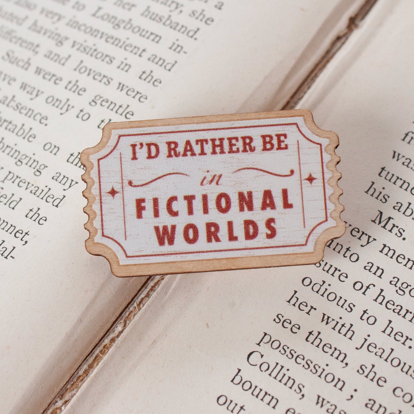 A die-cut vintage-style enamel pin with the text 'I'D RATHER BE in FICTIONAL WORLDS' in red lettering on a cream background, styled on top of an open book page.