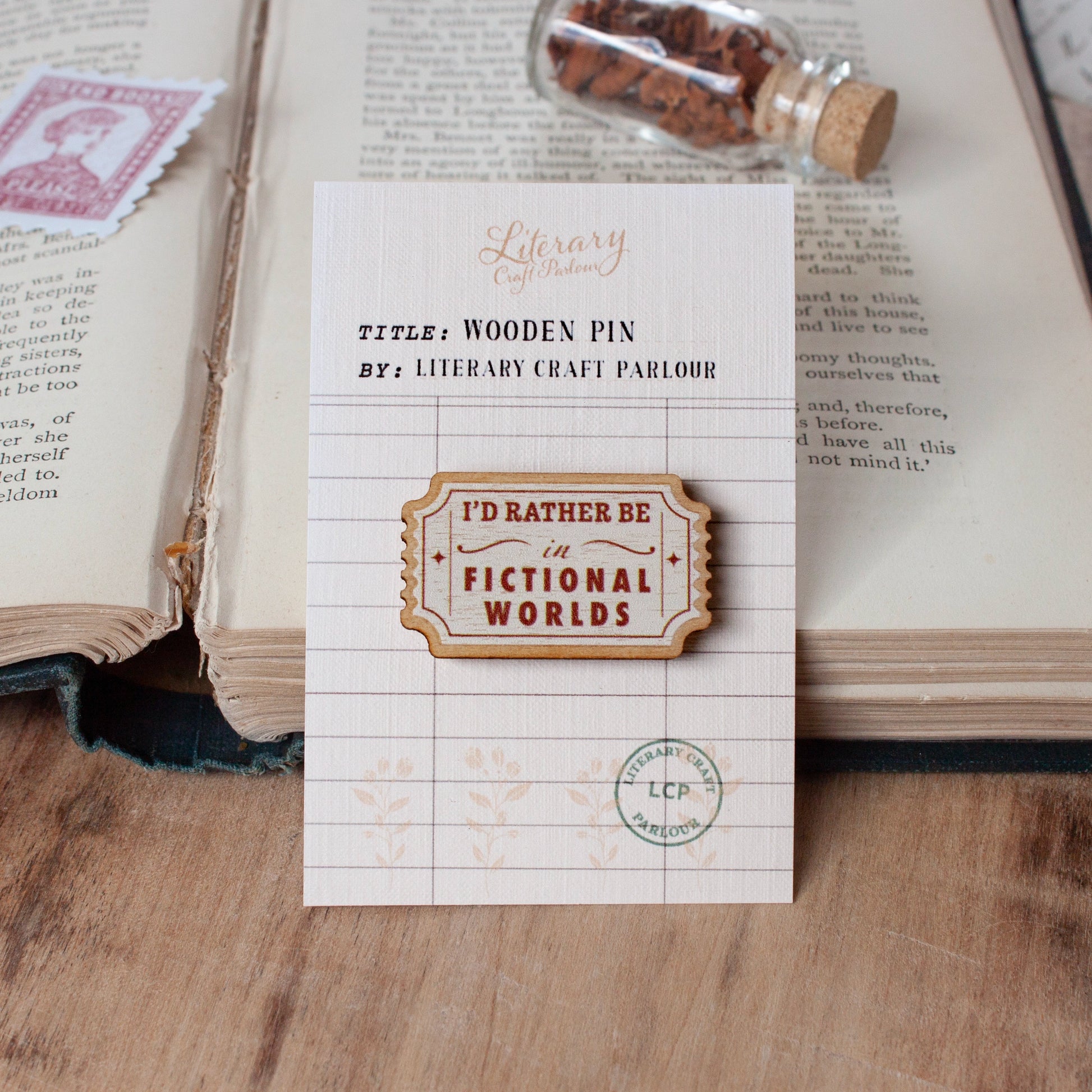 A wooden pin badge with the text 'I'D RATHER BE IN FICTIONAL WORLDS' displayed on its backing card, which is styled on a wooden surface next to an open book.