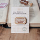 A wooden pin badge with the text 'I'D RATHER BE IN FICTIONAL WORLDS' displayed on its backing card, which is styled on a wooden surface next to an open book.