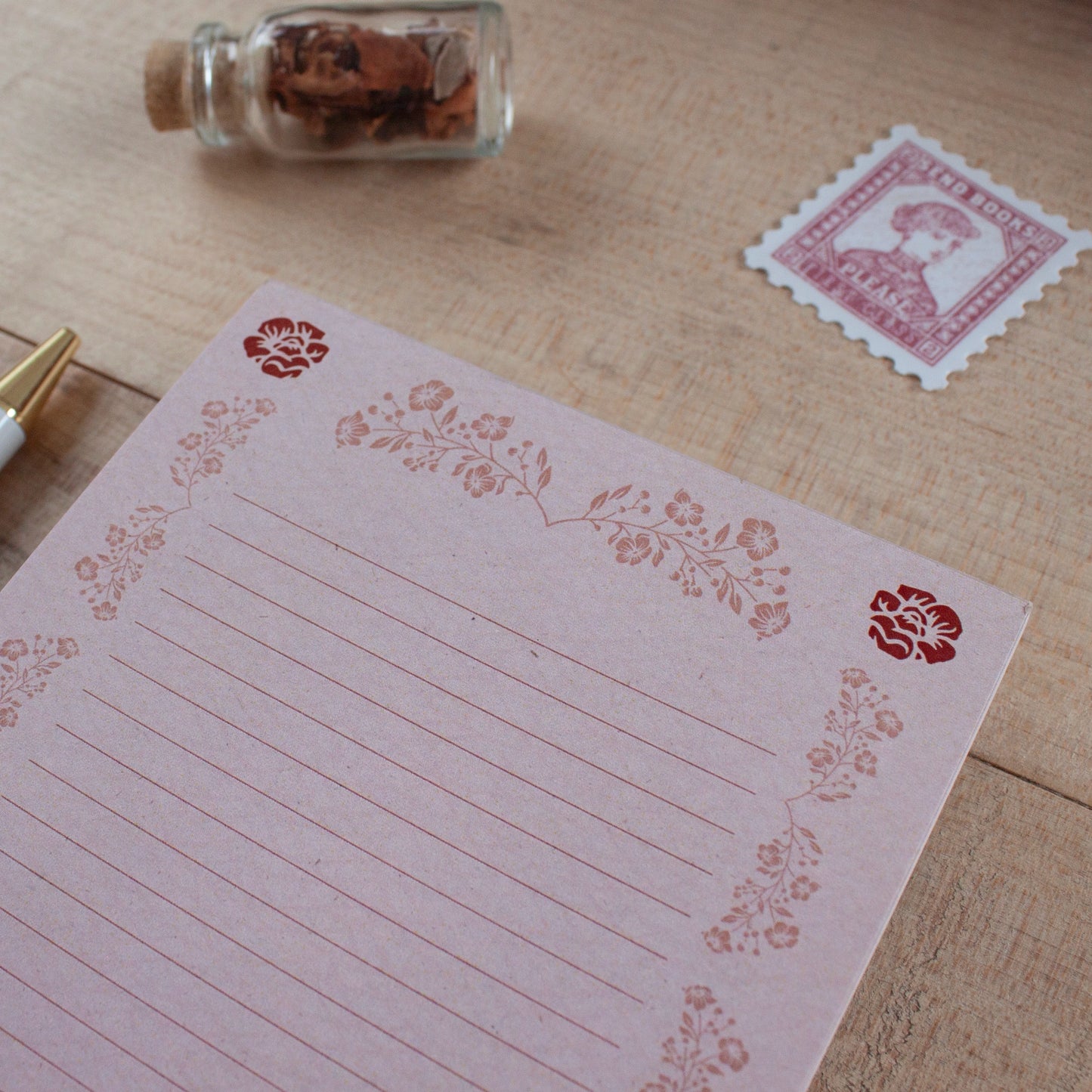 A pink lined notepad with floral border stamps, styled on a wooden desk next to a vintage stamp, a small glass bottle filled with flowers, and a gold pen.