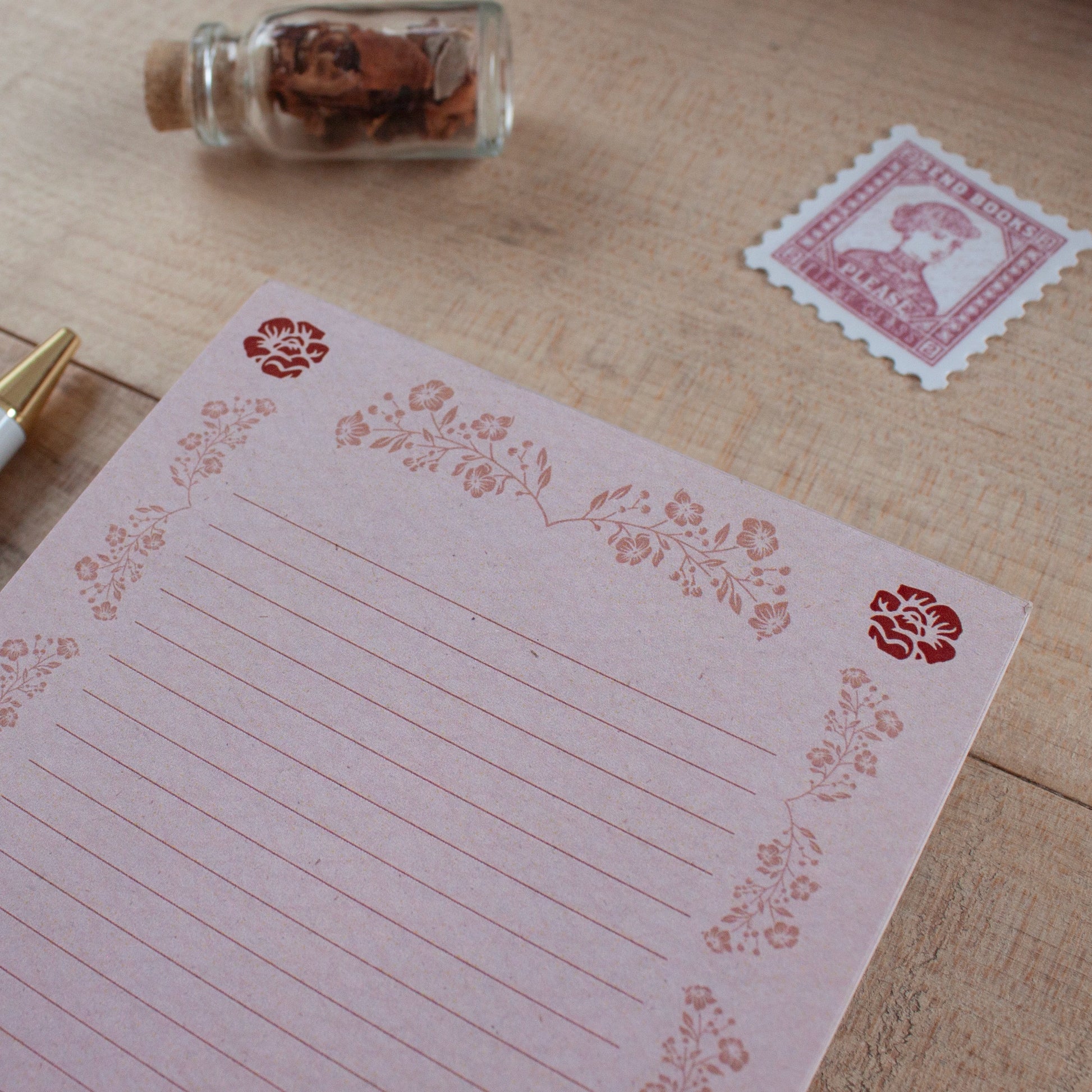 A pink lined notepad with floral border stamps, styled on a wooden desk next to a vintage stamp, a small glass bottle filled with flowers, and a gold pen.