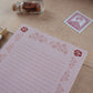 A pink lined notepad with floral border stamps, styled on a wooden desk next to a vintage stamp, a small glass bottle filled with flowers, and a gold pen.