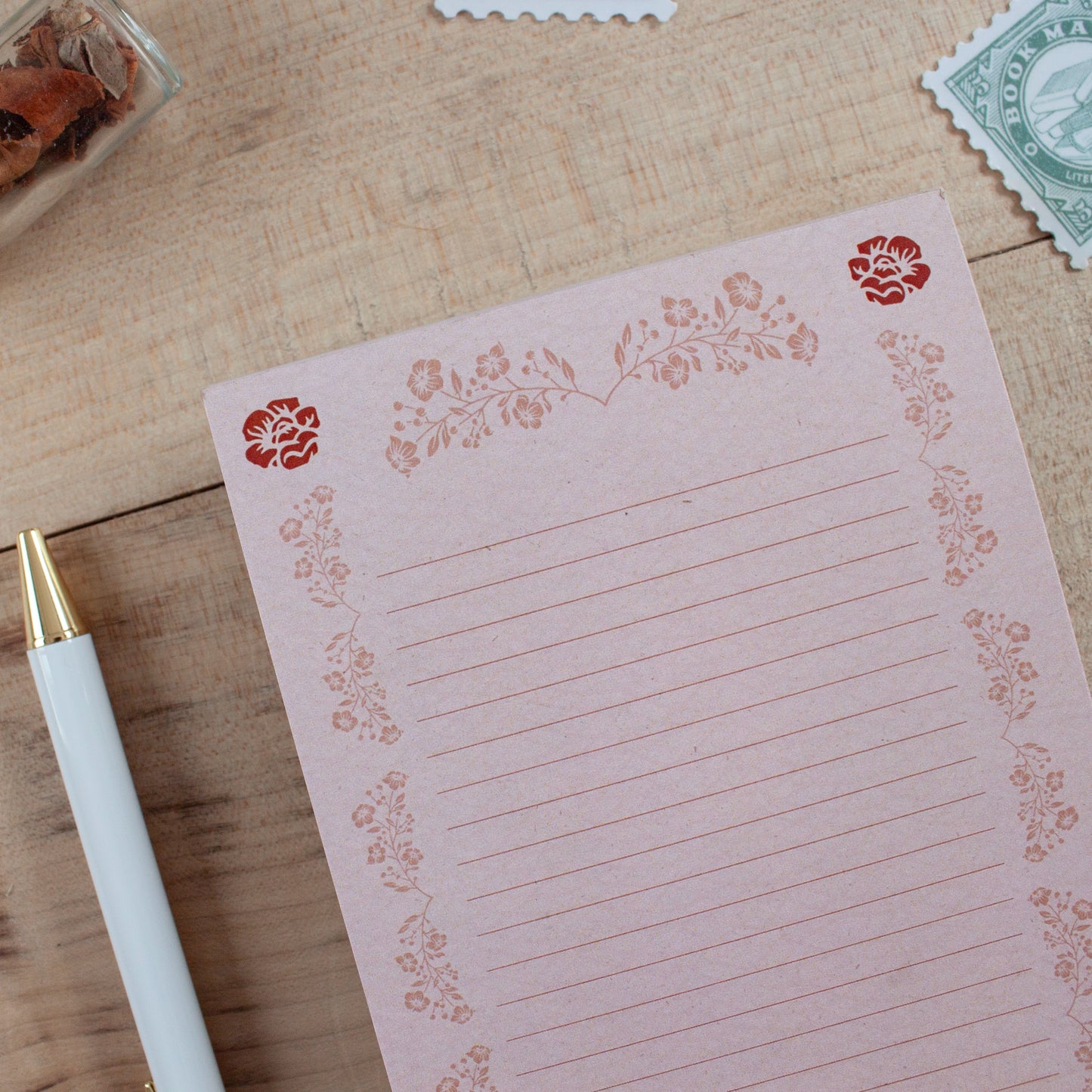 A pink lined notepad with a floral border design, styled on a wooden tabletop next to a white pen.