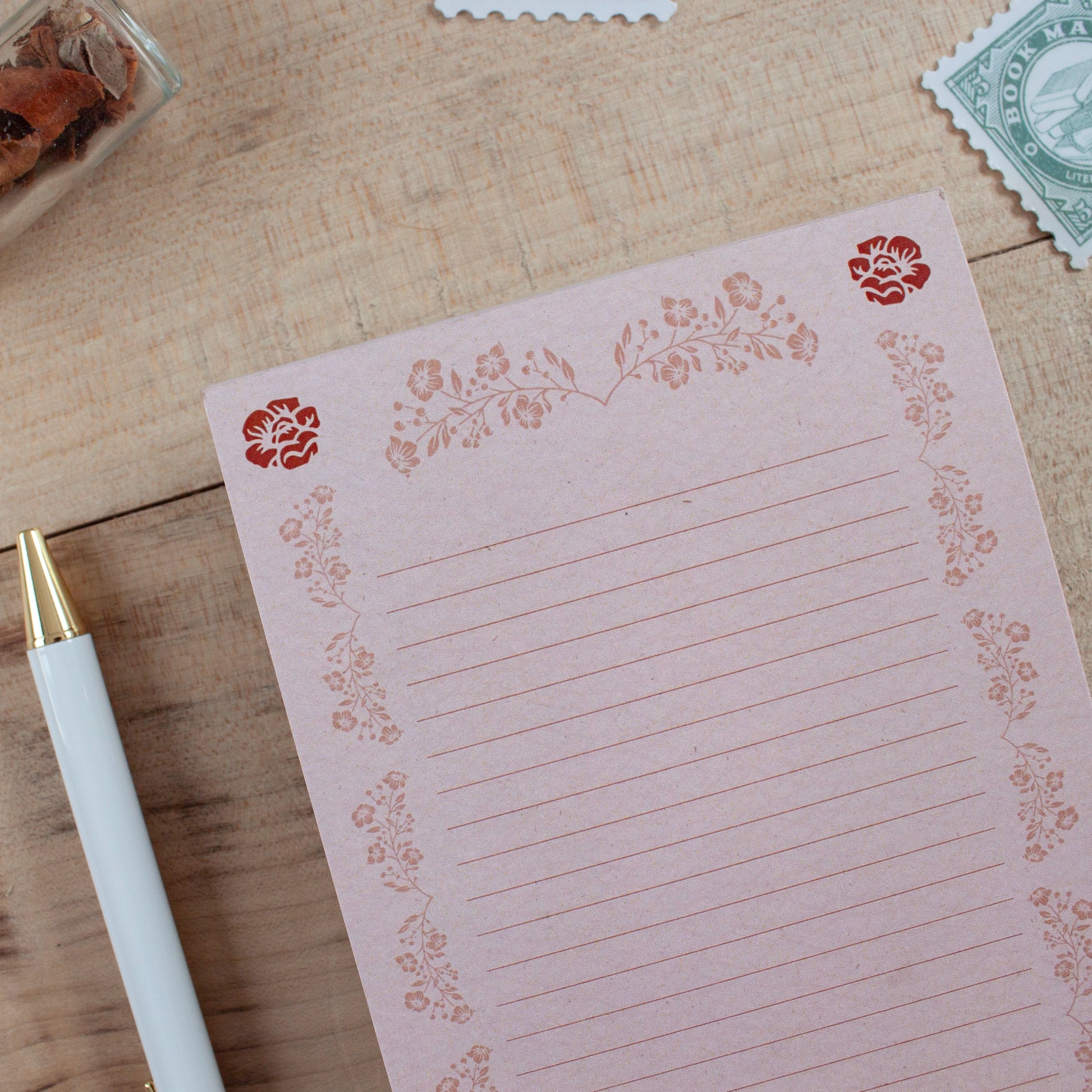 A pink lined notepad with a floral border design, styled on a wooden tabletop next to a white pen.