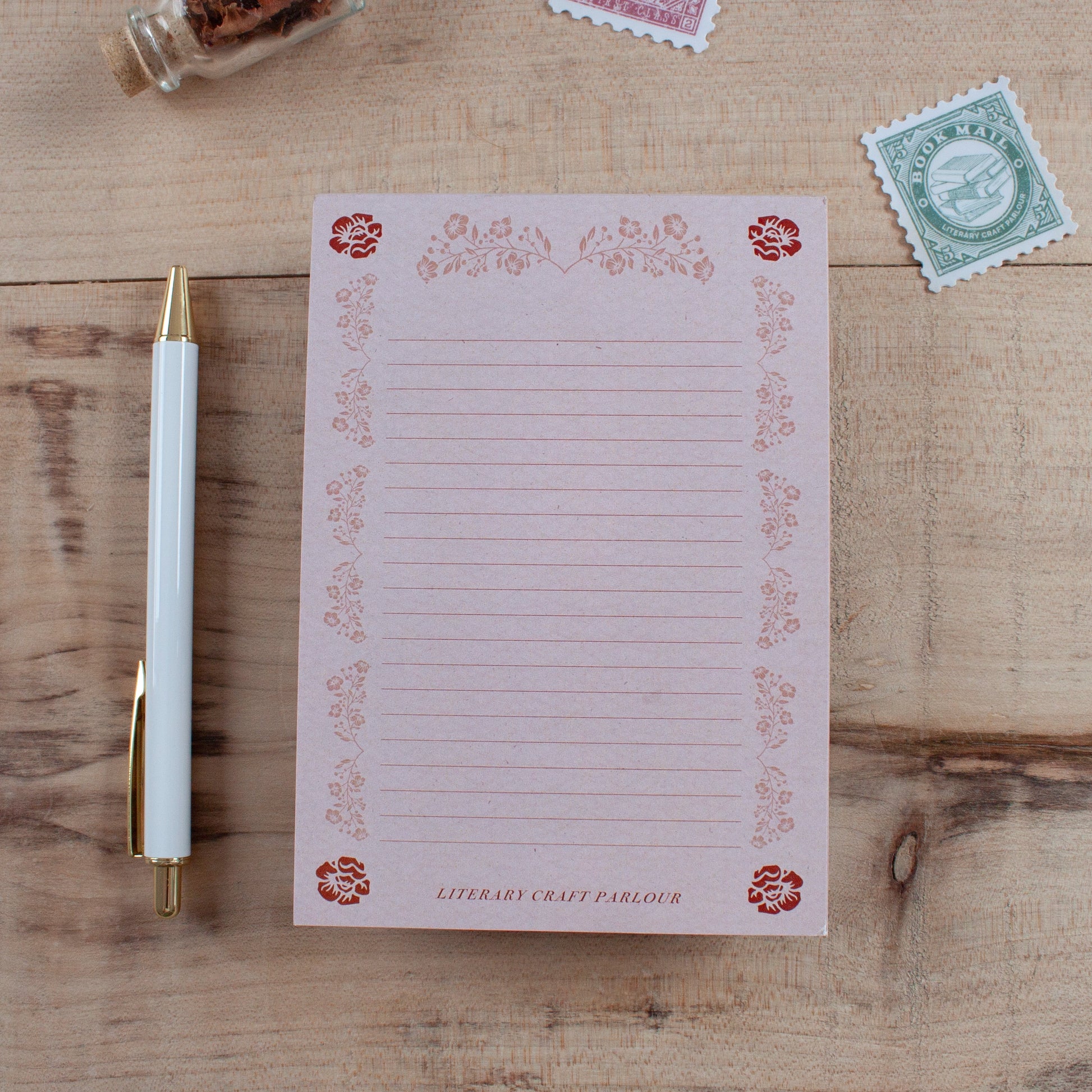 A pink lined notepad with a decorative floral border and the text 'LITERARY CRAFT PARLOUR' at the bottom, styled on a wooden tabletop with a white pen and postage stamps nearby.
