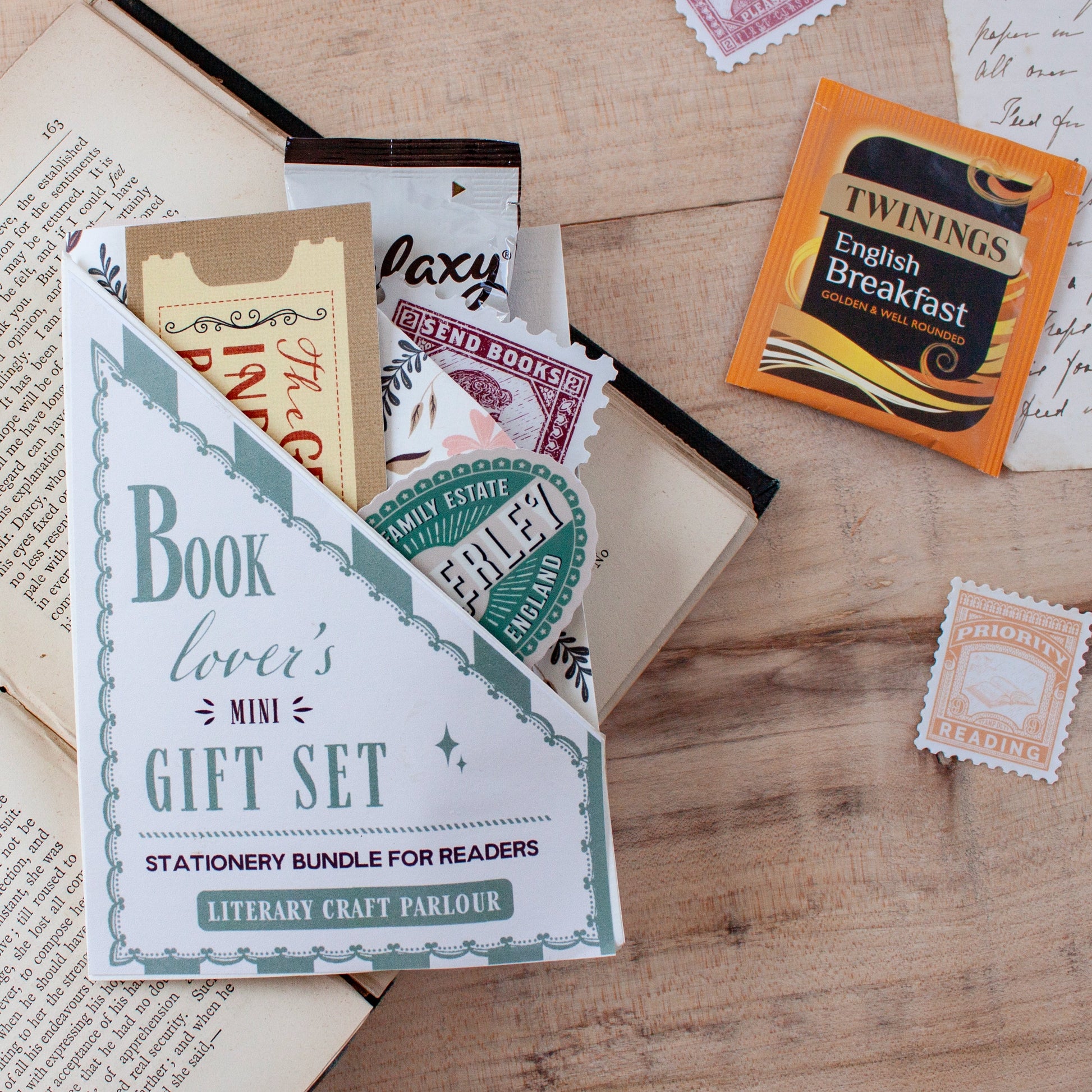 A book-themed mini gift set displayed on a wooden tabletop, featuring a white envelope-style holder labeled 'Book lover's Mini Gift Set' with stationery pieces peeking out, alongside a Twinings English Breakfast tea packet a Galaxy hot chocolate sachet and vintage-style postage stamps.