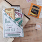A book-themed mini gift set displayed on a wooden tabletop, featuring a white envelope-style holder labeled 'Book lover's Mini Gift Set' with stationery pieces peeking out, alongside a Twinings English Breakfast tea packet a Galaxy hot chocolate sachet and vintage-style postage stamps.