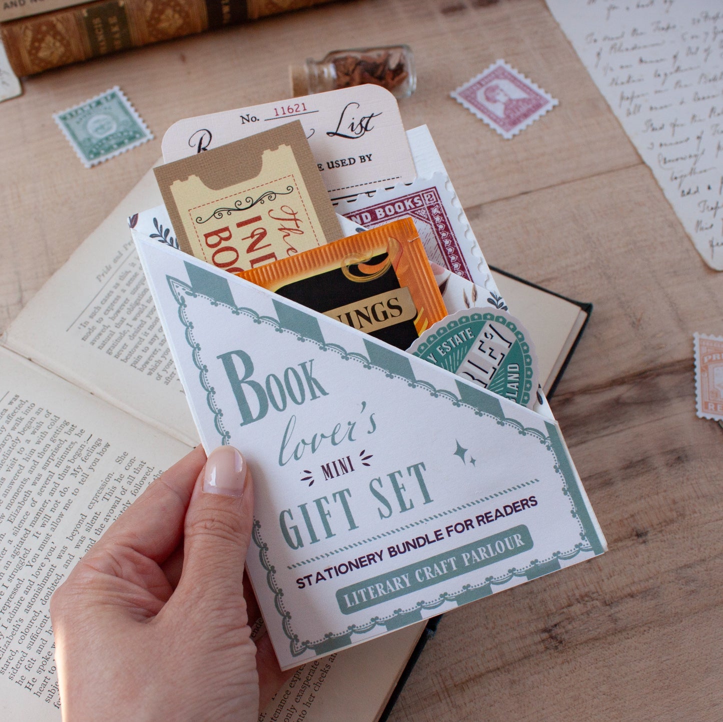 A hand holds a triangular shaped pocket labeled 'Book lover's mini gift set' open to reveal stationery and small literary-themed craft items tucked inside, photographed on a wooden tabletop with books and stamps around it.