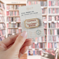 A hand holds a small wooden pin mounted on a branded backing card. The pin is shaped like a vintage ticket with the text 'I'D RATHER BE IN FICTIONAL WORLDS' in red lettering.